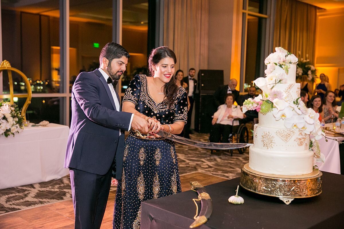 Sheikh Groom wearing a navy suit and Indian bride wearing a navy and gold saree cut their white wedding cake with gold mandala details and white orchids with a sword.