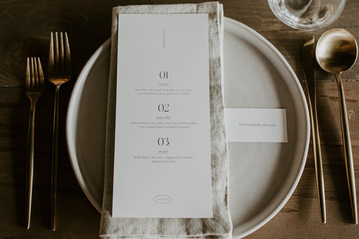 A white menu card with black font atop a linen napkin and white plate set on a wooden table with gold silverware.