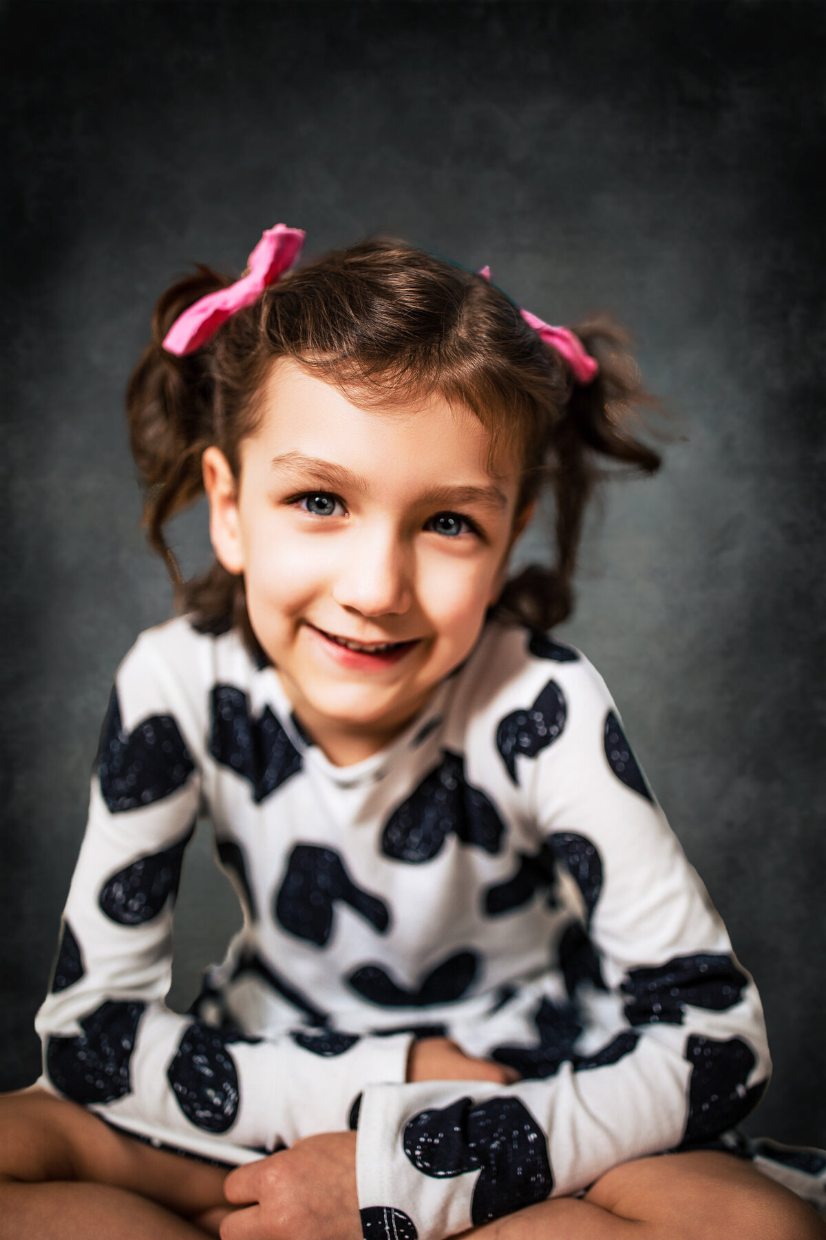 A girl with pink bows and a black and white top poses on a gray Studio background for larger than life photography at the studio
