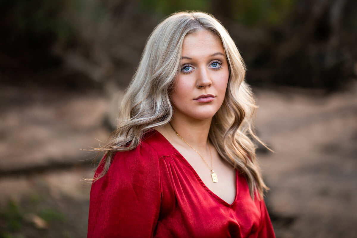 A stunning close up portrait of a beautiful blonde haired blue eyed teen girl wearing a red dress standing on the lake shore. Captured by senior photographer Dynae Levingston.