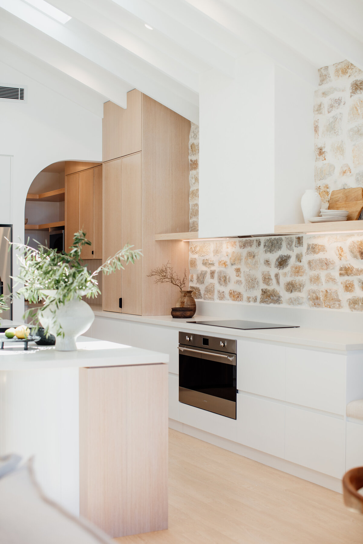 This modern kitchen features a stone wall, wooden cabinets, white countertops, and a built-in oven, exemplifying luxury home design in Newcastle. A vase with greenery graces the island, adding a touch of nature to this carefully curated space by Newcastle kitchen and bathroom designers.
