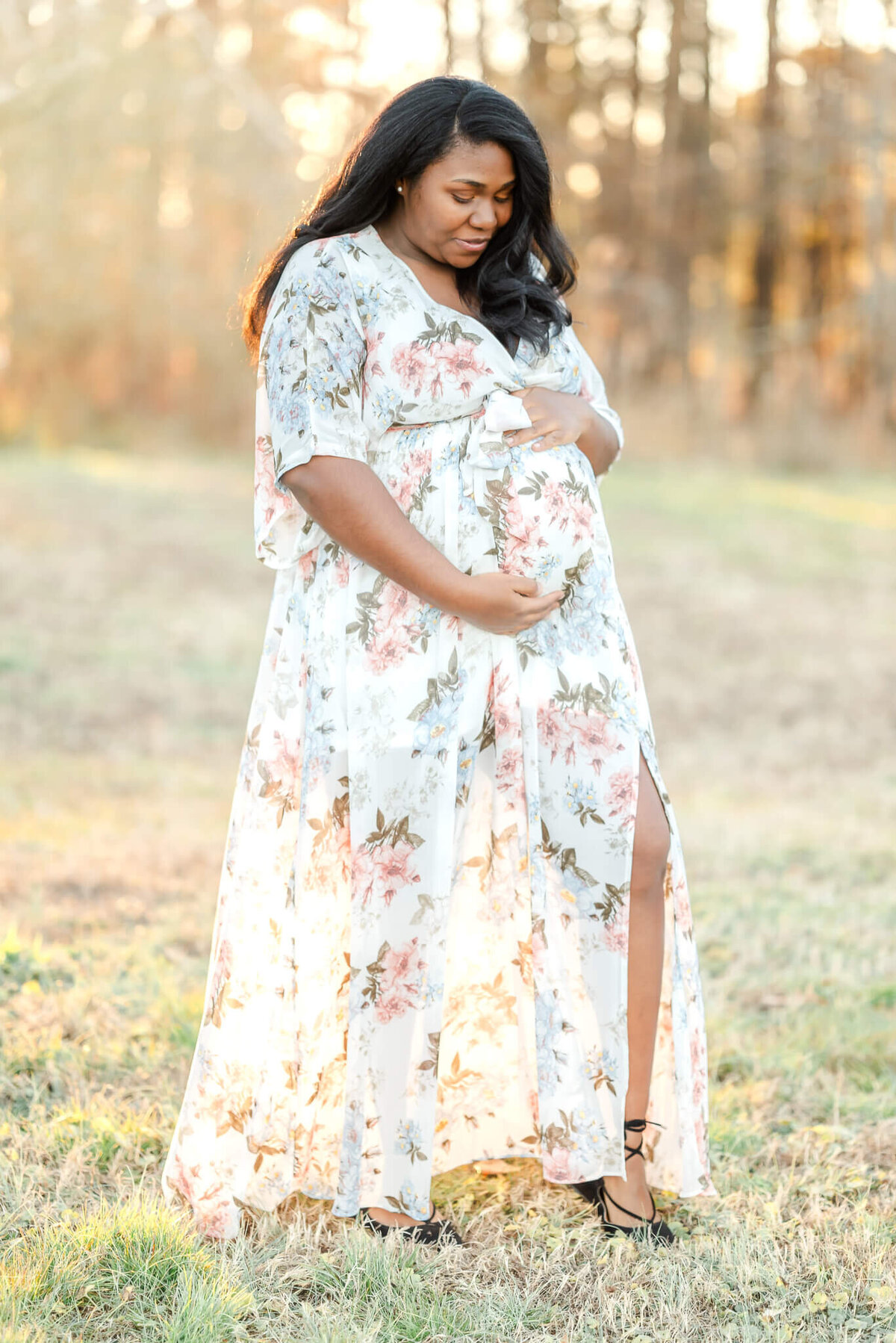 A pregnant woman, wearing a white floral dress, holds and looks down at her belly during her Chesapeake maternity photography session.