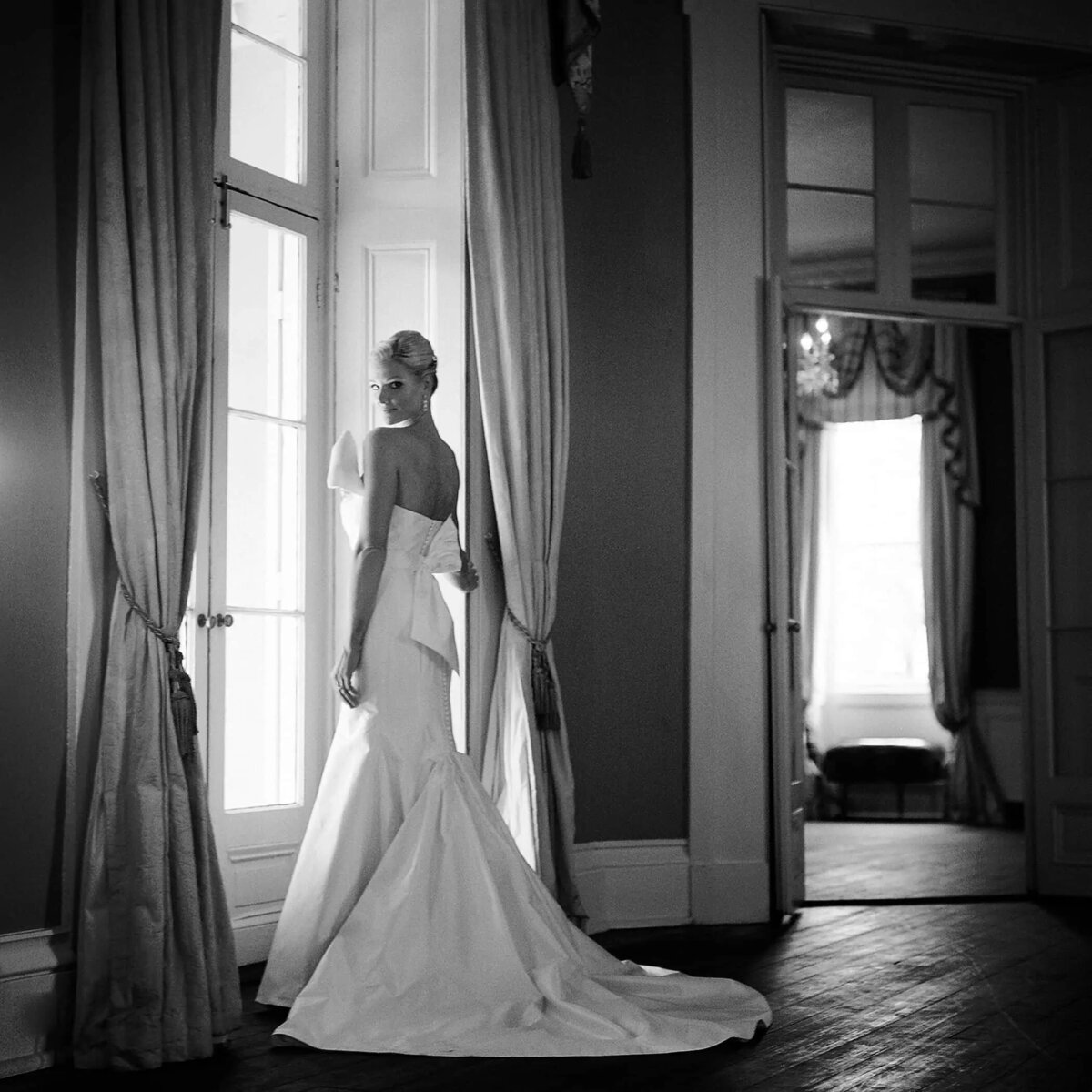 A bride in a chic wedding dress stands by a window in a vintage room, her back to the camera
