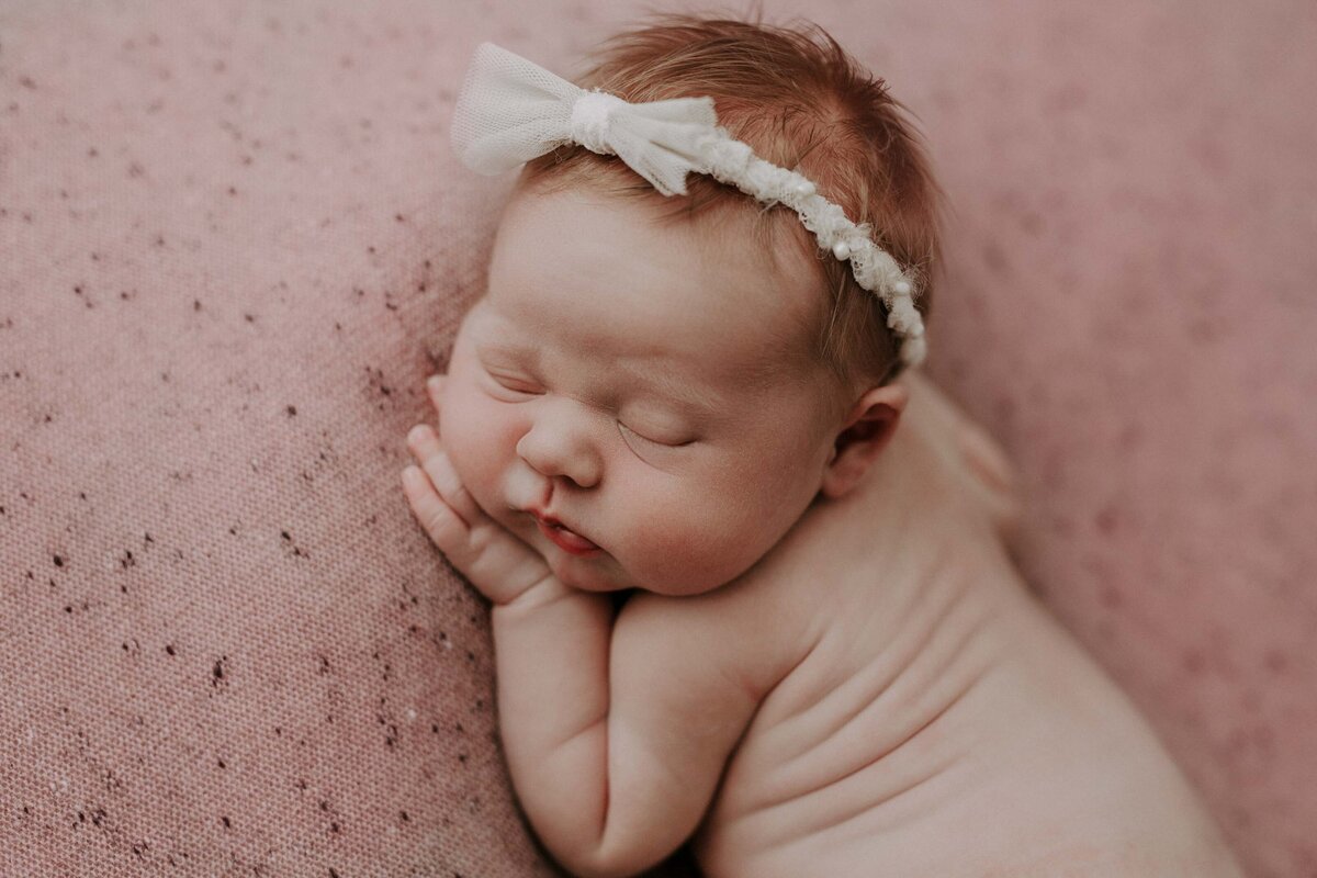 Studio newborn photography - Baby resting on a pink eggshell blanket. Baby is bare wearing only a delicate cream bow headband. Baby's hand is resting under her cheek.