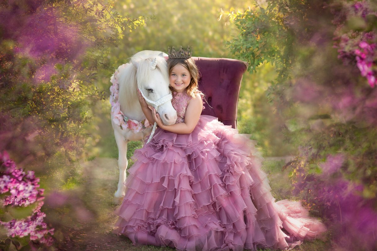 girl-in-coutuire-dress-sitting=-on-a-chair-hugging-a-unicorn-with-pink=flowers-surround