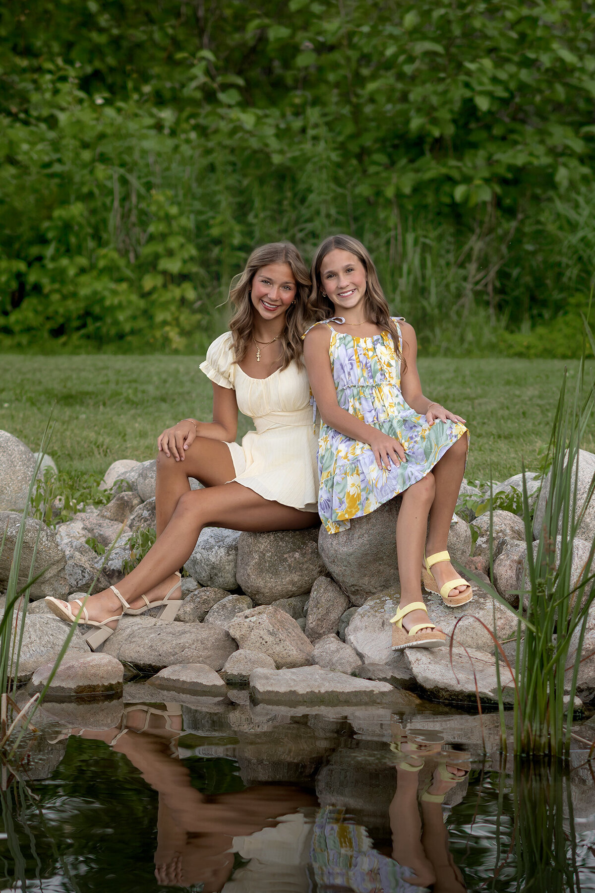 2 sisters lean against each other as they sit next to a pond