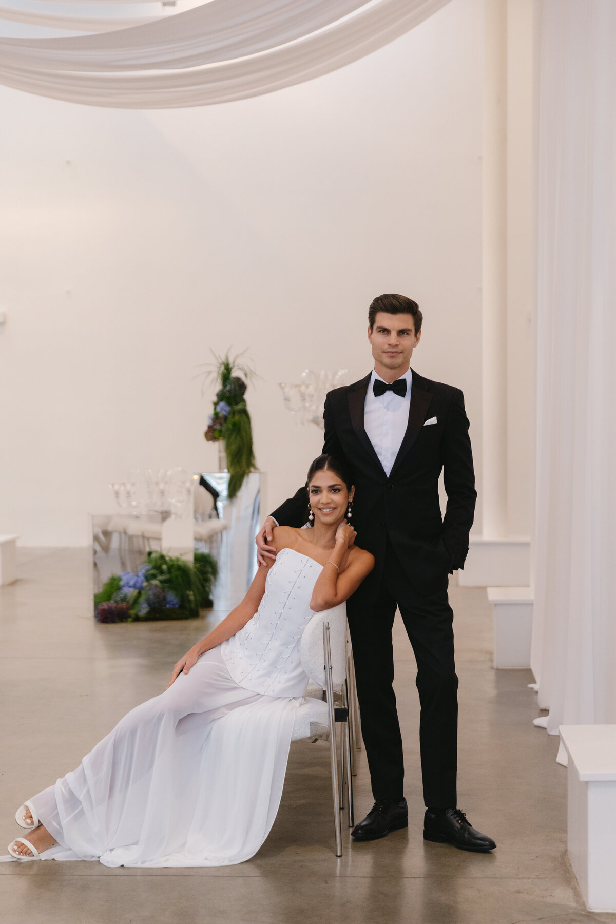 Bride and groom posed together in an elegant modern wedding editorial with white draping and floral installations.
