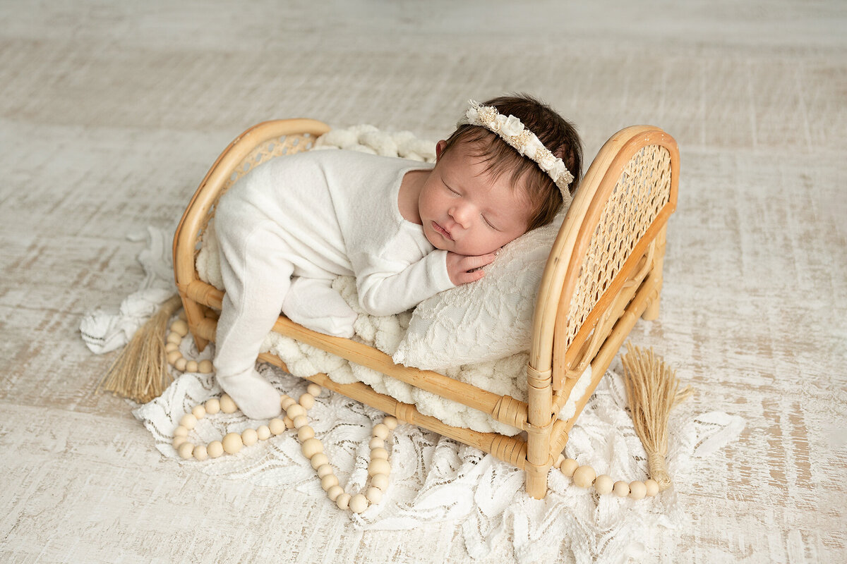 columbus-ohio-newborn-photographer-baby-girl-in-white-footed-romper-white-headband-laying-on-tummy-on-rattan-doll-sized-bed