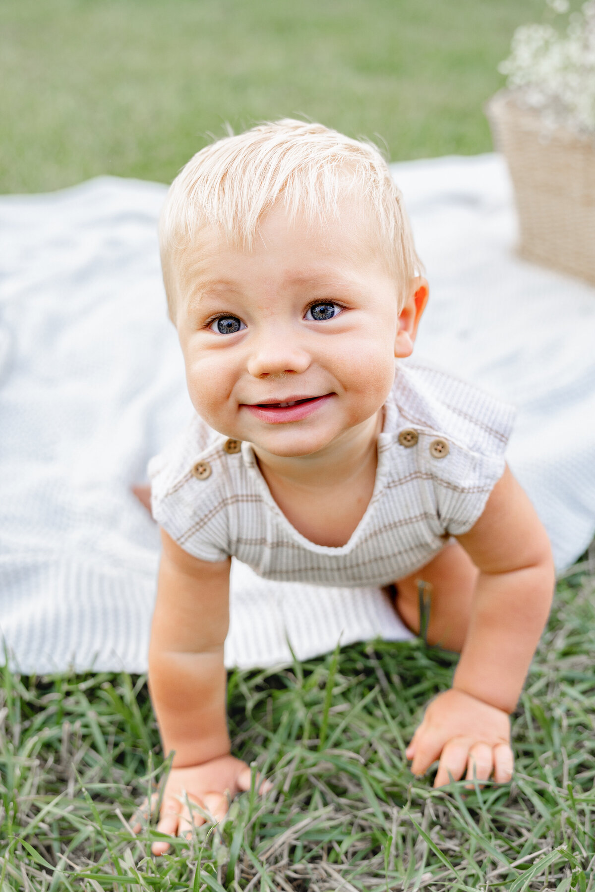 A cheerful blonde-haired baby crawls on the lush green grass, his bright blue eyes sparkling with excitement in an Eagle Mountain field.