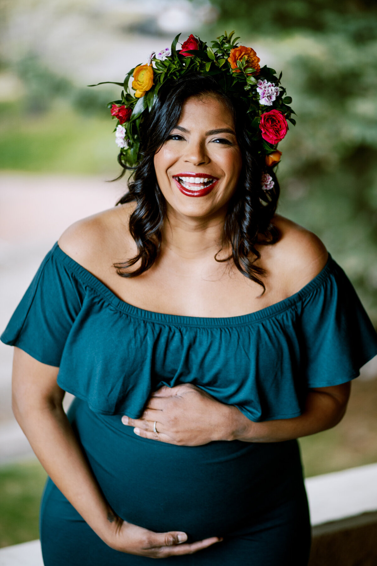 beautiful smiling woman in floral wreath and blue dress laughs while holding baby bump in boulder's chautauqua park