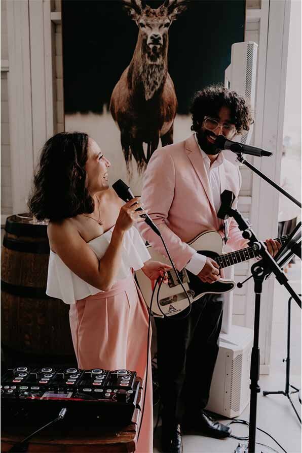 Acoustic duo dressed in coordinated blush outfits singing and playing guitar at a wedding venue.