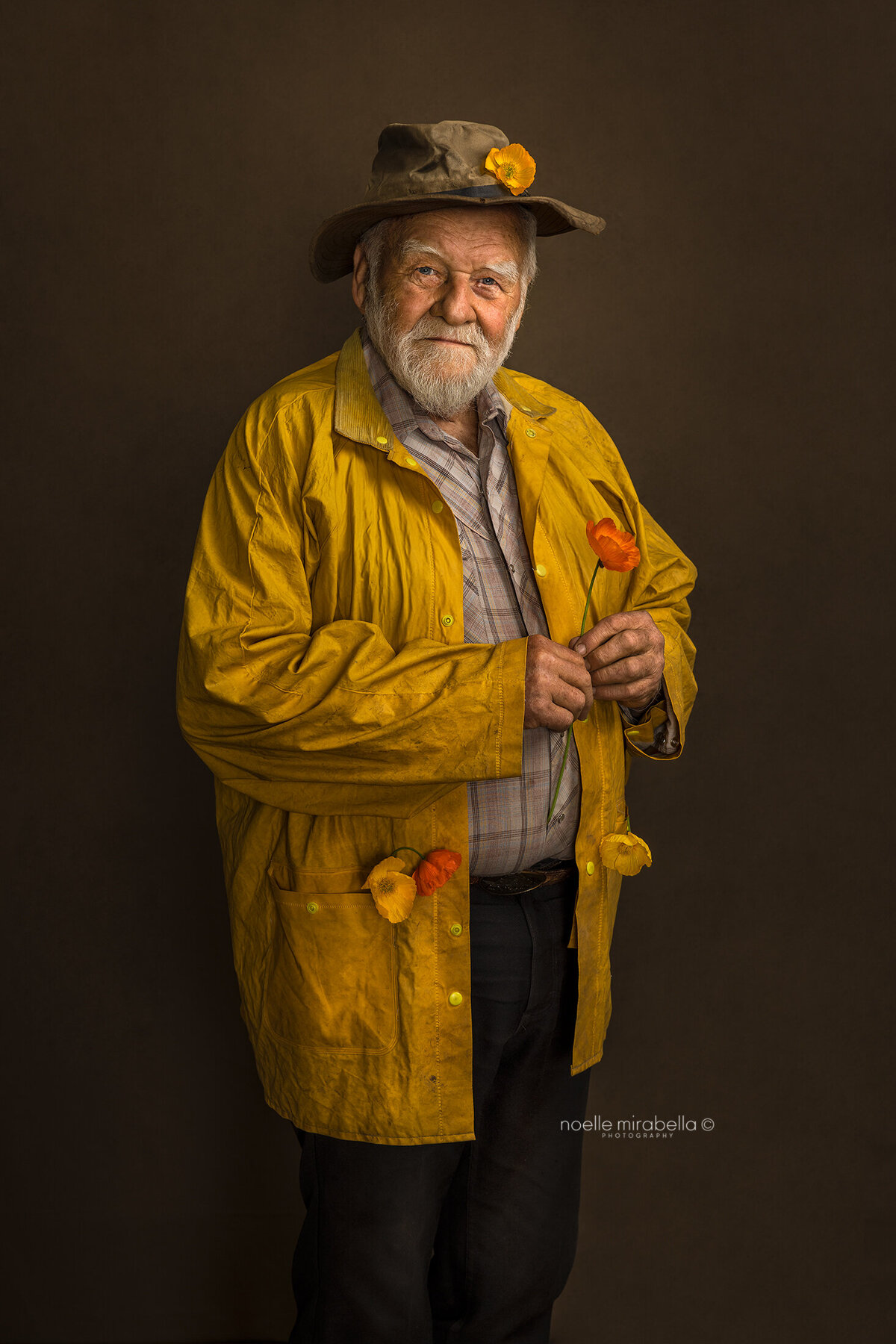 Elderly man dressed in a plaid shirt, leather hat, and yellow raincoat holding an Icelandic poppy. 