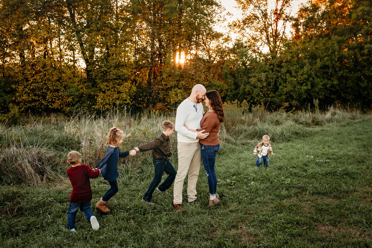 A couple embraces in a grassy field while their four children play joyfully, hand in hand. The sun sets, casting a warm glow reminiscent of timeless outdoor portraits captured by a Bloomington family photographer. Trees frame this moment of love and laughter.