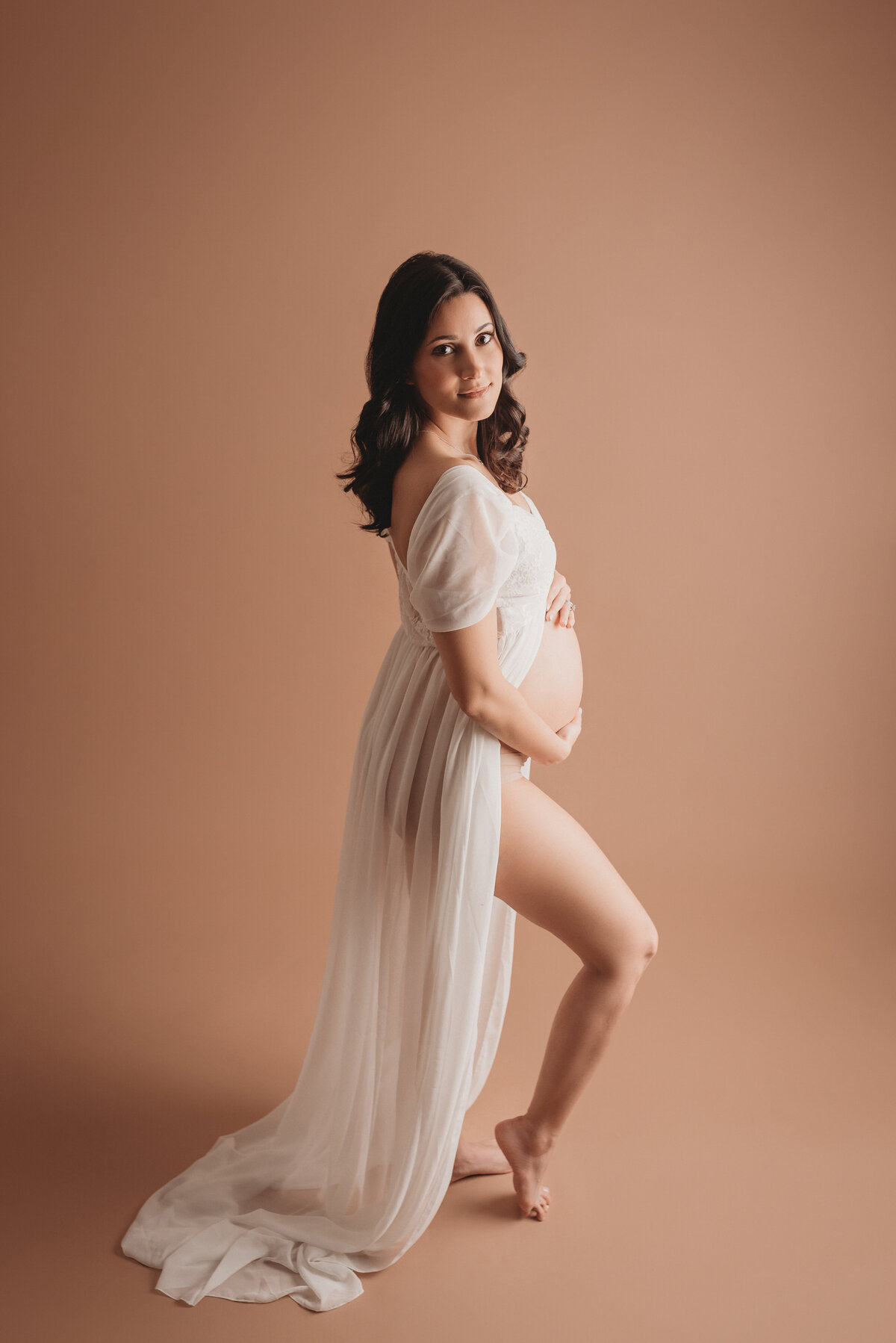 Pregnancy portrait on tan backdrop with 36 week pregnant woman holding baby bump looking at the camera and wearing sheer white maternity gown open on tummy