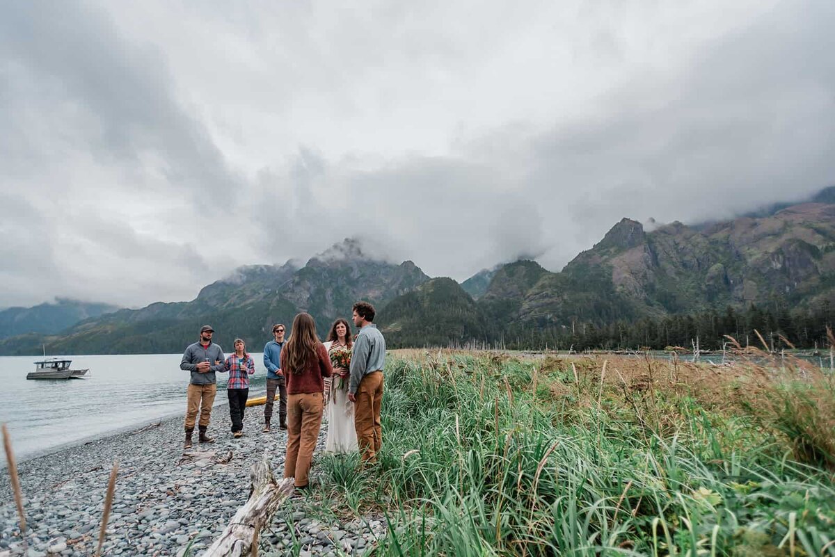 A bride and groom in formal wedding clothes and their guests are standing on a pebble covered beach during their adventure luxe elopement in Kenai Fjords.  They are tiny in the frame and behind them, a huge, hulking mountain of stone and lush green trees is shrouded in mist, so the top is hidden.  Their private boat taxi that brought them too the island is visible small in the distance out on the water.  It feels like Jurassic Park..