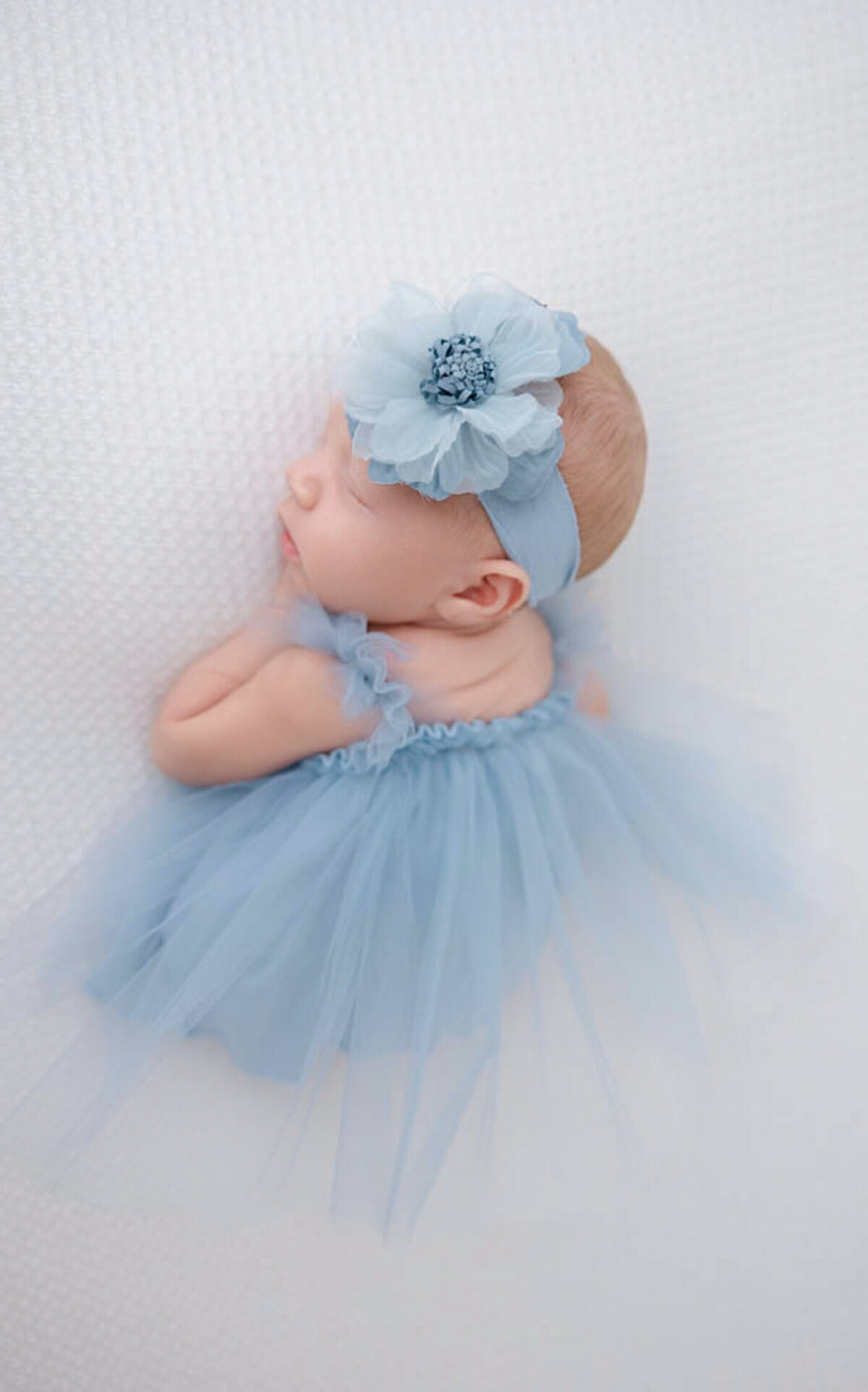 Infant girl wearing a blue frilly dressed and blue flowered headband and lying on a white blanket.