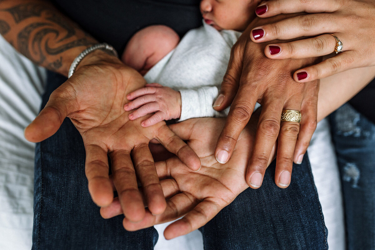 dad with tattoos hands next to mom's and baby's while resting on dad's lap