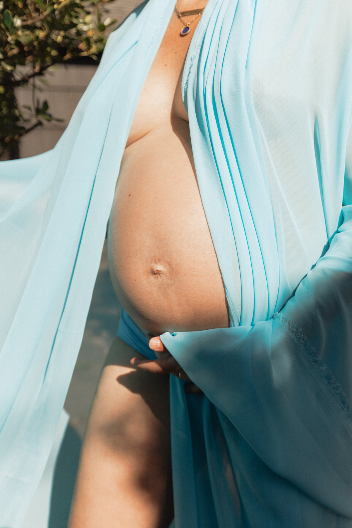 Pregnant woman draped in sheer blue fabric, in the sunshine during maternity photoshoot