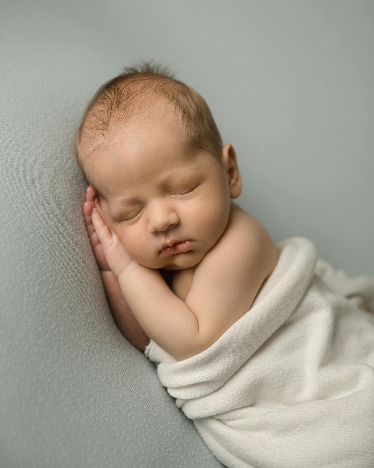 Newborn boy sleeping with his head resting on this two hands.  Wrapped in white with a light blue background.