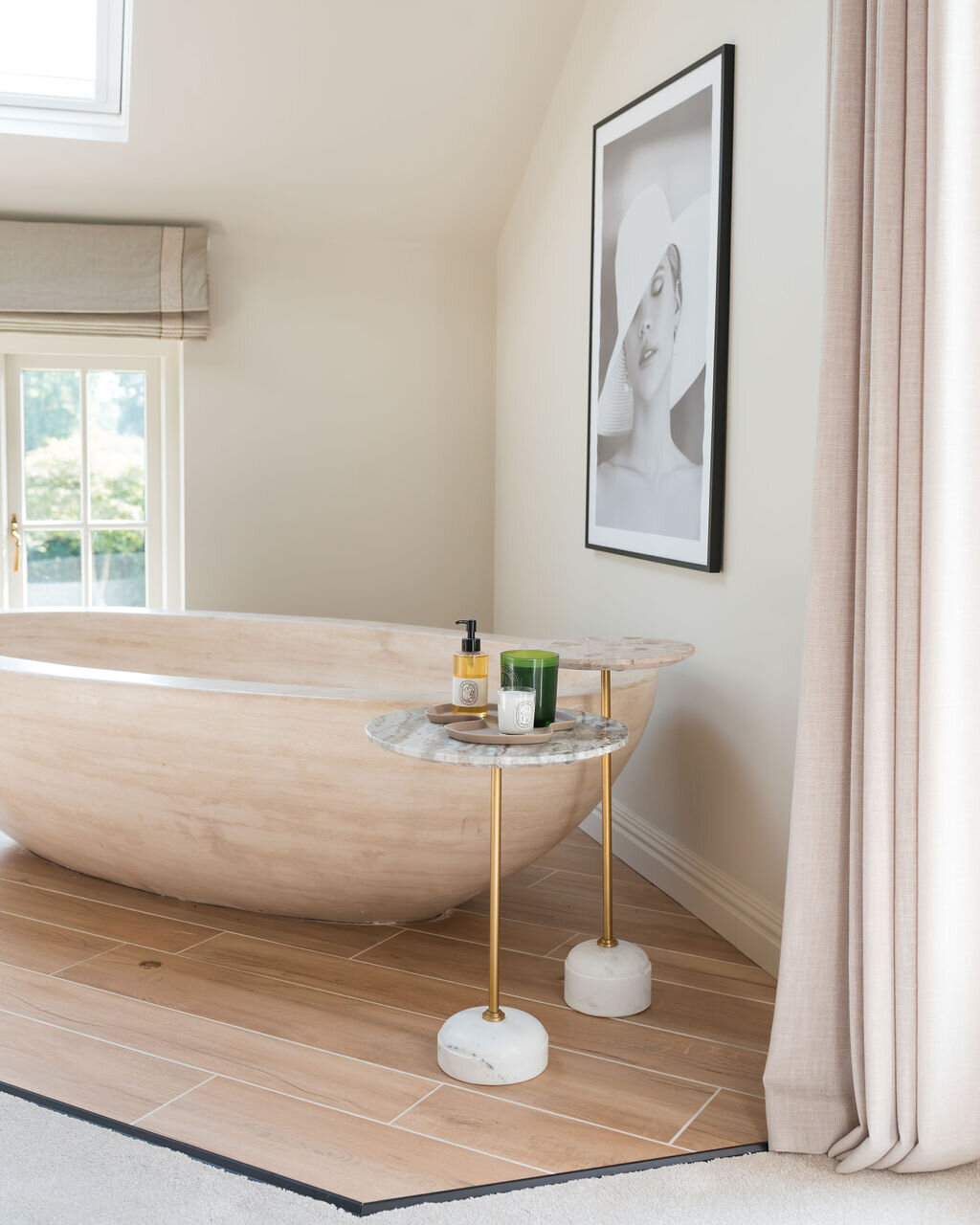 A minimalist neutral bedroom features a large, freestanding wooden bathtub under a skylight. There's a small side table with decor and a framed black-and-white photo on the wall. The space is bright with natural light and neutral tones.