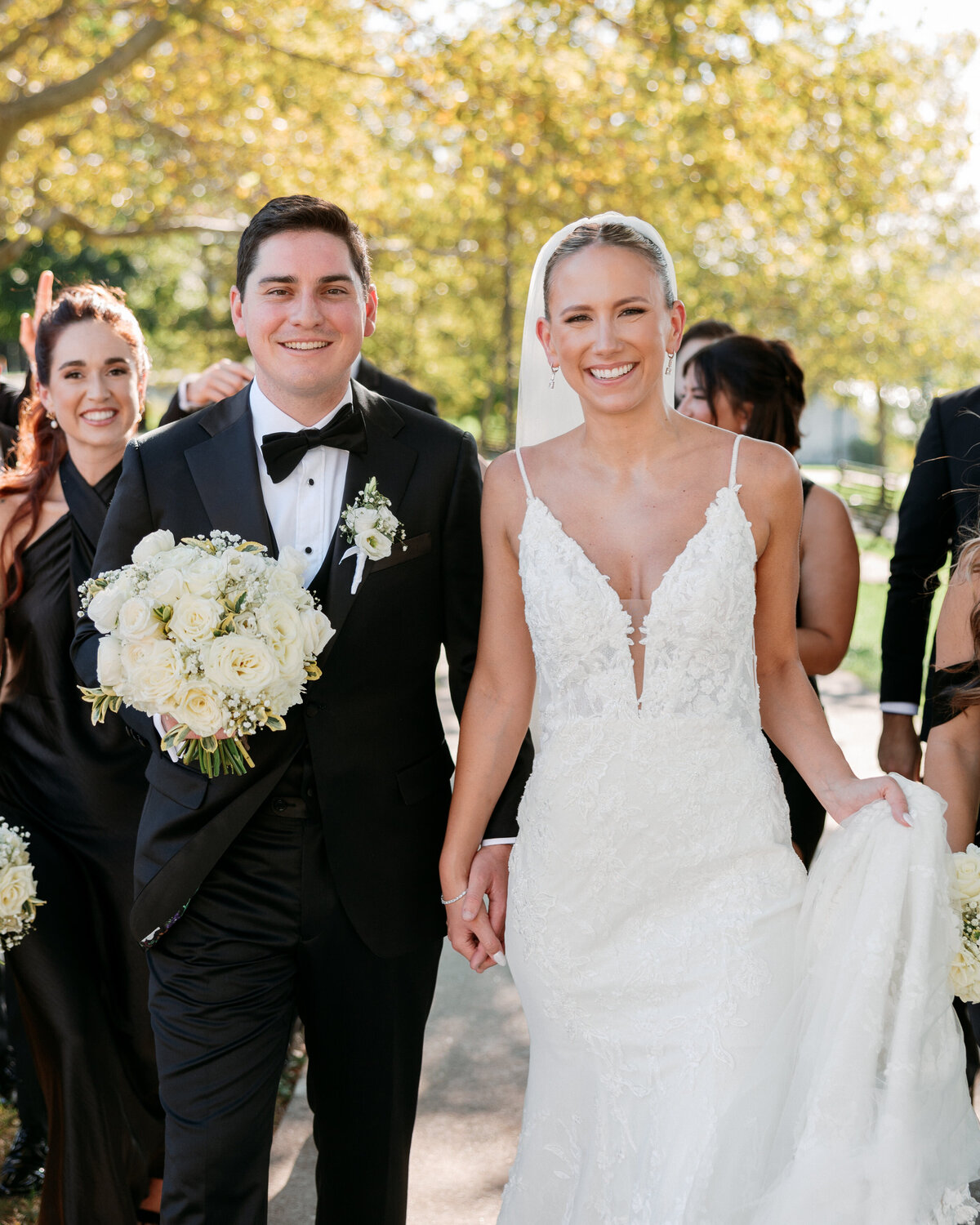 Radiant couple walks hand in hand with their wedding party in a sunny park, smiling and joyful, beautifully documented by a Philadelphia wedding photographer.