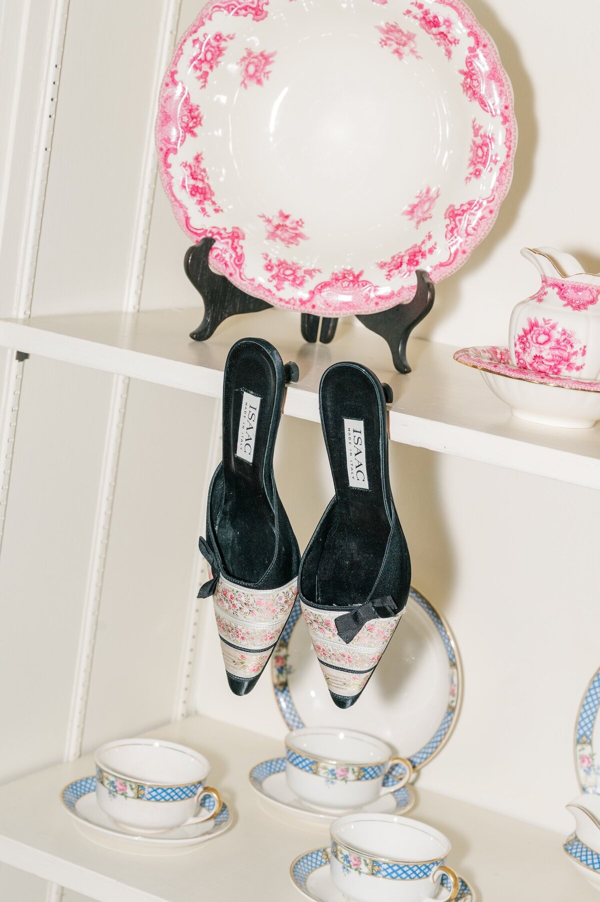 A pair of black and pink shoes hanging from a shelf that is holding teacups and saucers. 