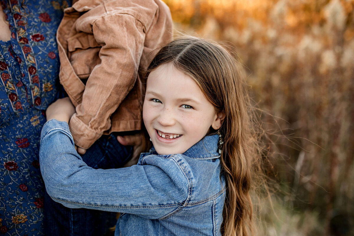 A young girl with long brown hair wears a denim jacket and smiles at the camera. She holds a smaller child in a brown jacket. They are outdoors with tall grass and blurred autumn colors in the background.