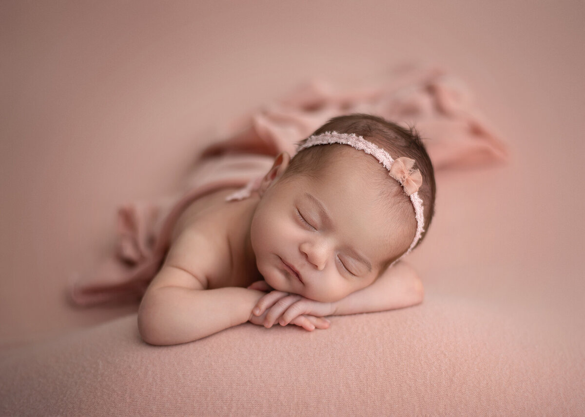 newborn with head on hands and pink headband and blanket