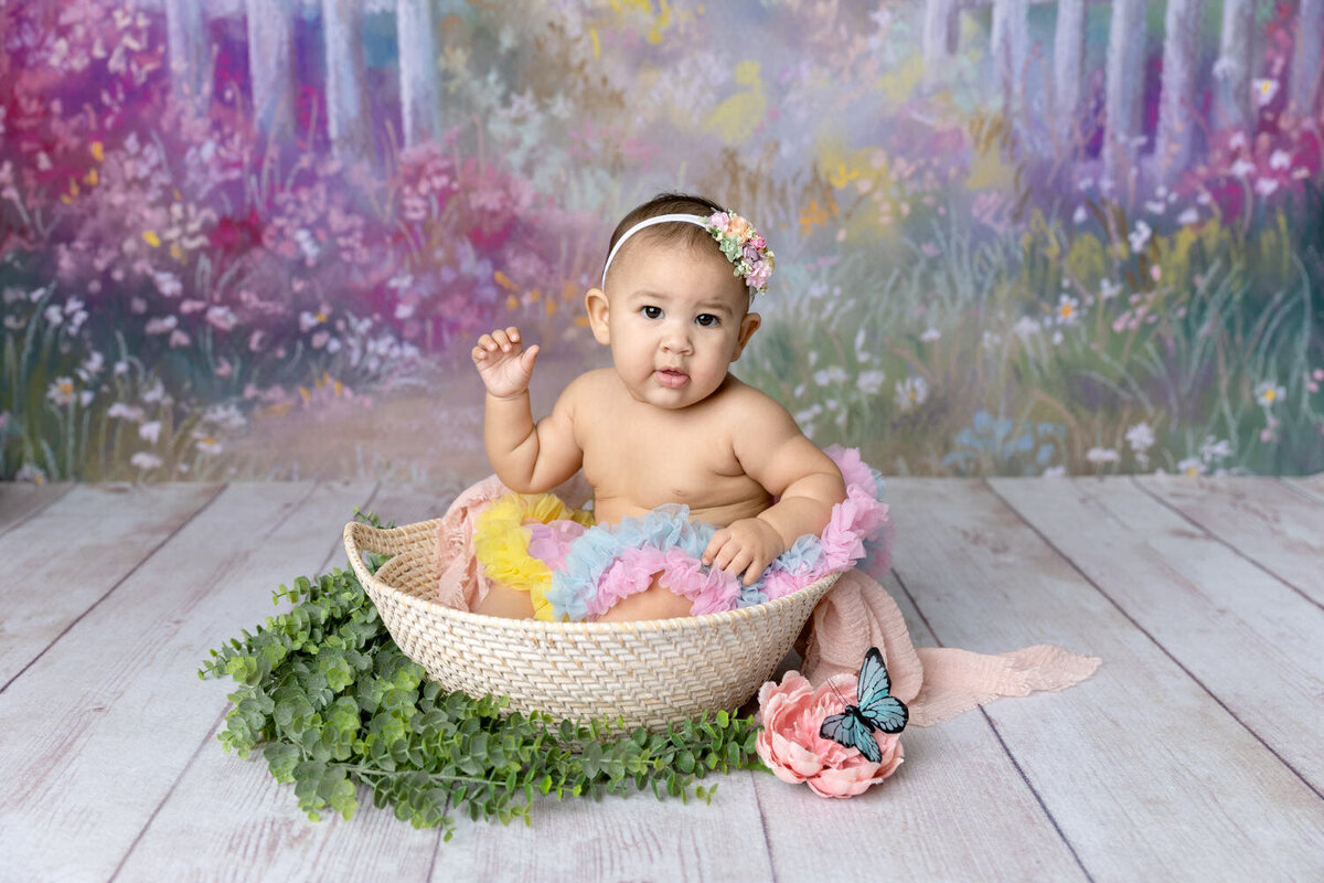 Baby girl sits in a basket in front of a floral background
