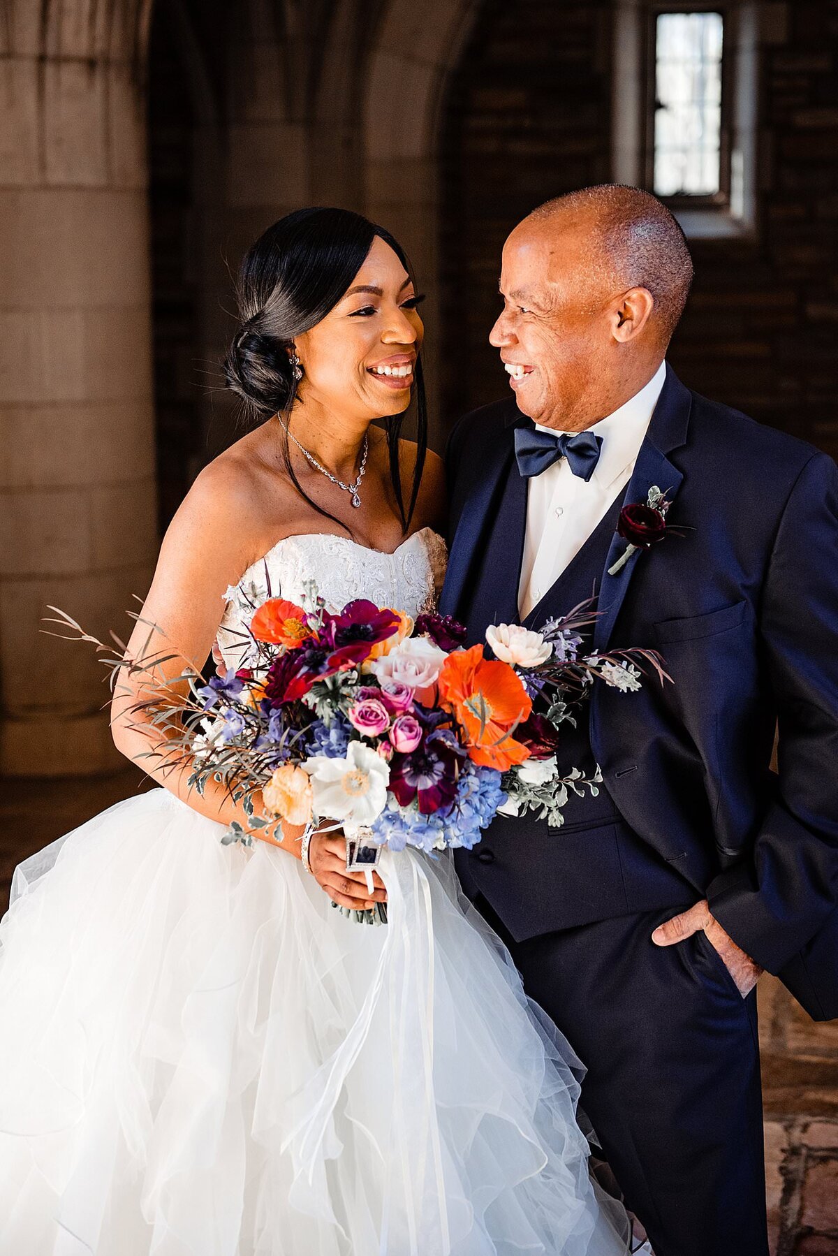 The bride and groom share a laugh inside the church. The groom is wearing a black tuxedo with a white shirt and a black bow tie. The bride is wearing a strapless gown with a sweetheart neckline and  ruffled organza skirt. She is holding a large colorful bouquet of flowers. The flowers are deep red, deep orange, blue, deep purple, yellow, blush and ivory.