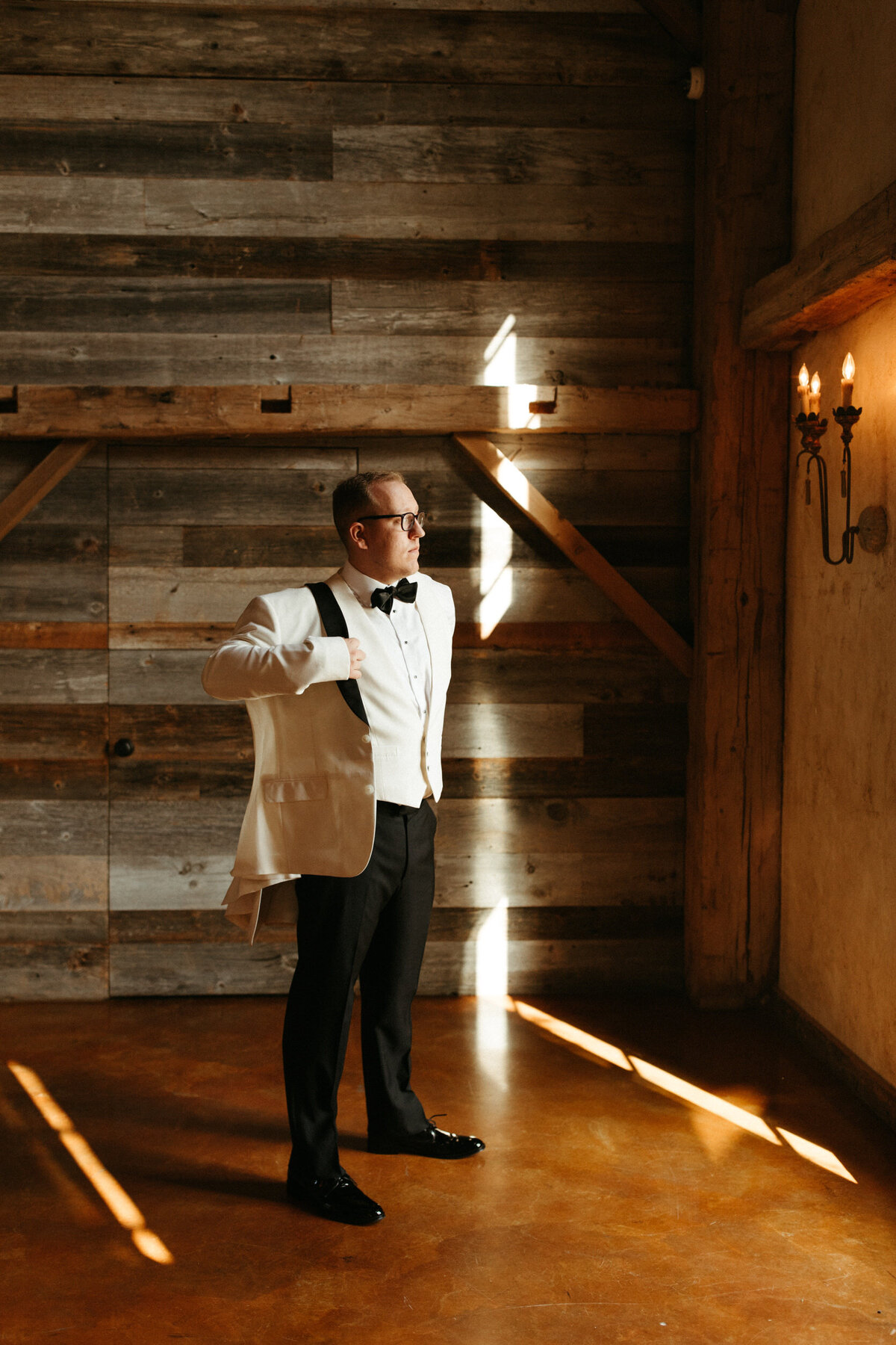 Groom wearing a bowtie putting his white tuxedo jacket on as he is getting dressed