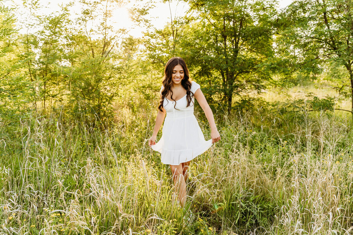stunning senior girl in a white dress walking through grasses, flowers and trees while smiling