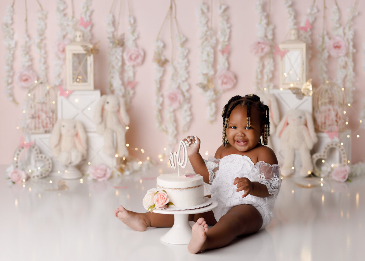 Somebody is one floral bunny cake smash in West Palm Beach and Loxahatchee Florida cake smash studio. Baby is wearing a white lace off the shoulder romper sitting behind a floral decorated white cake. In the background is a soft pink backdrop with hanging florals and plush bunny rabbits with lanterns and scattered flowers.