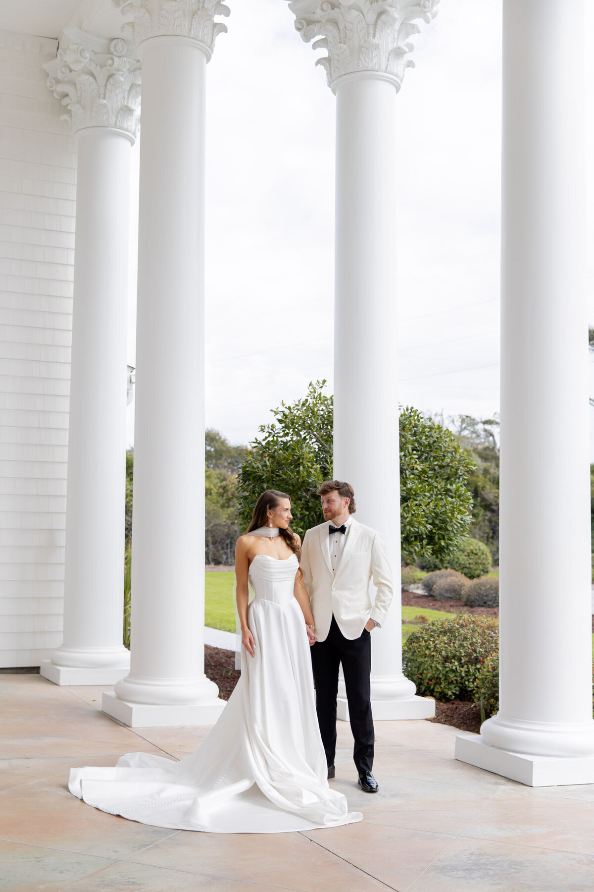 A bride and groom stand holding hands with a row of very tall white columns next to them.