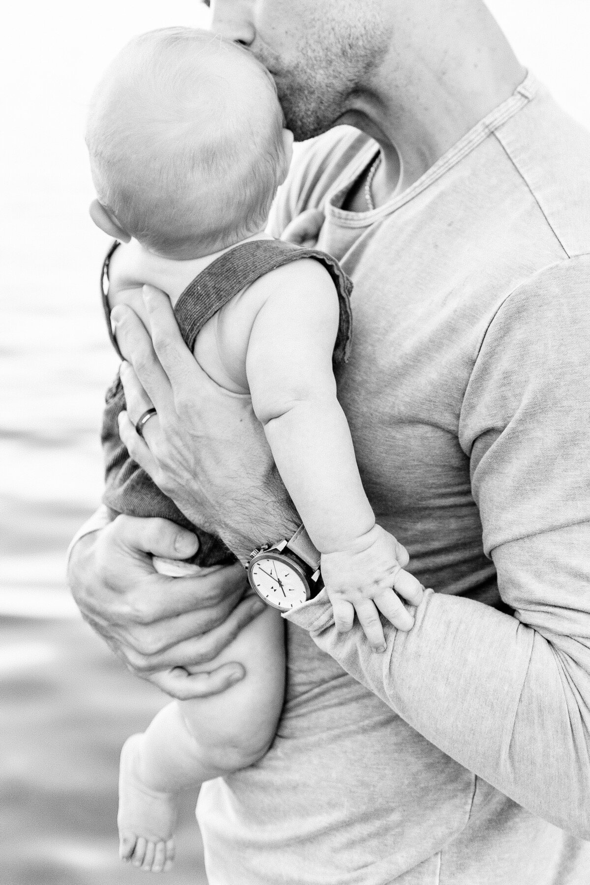 Black and white image of father holding and kissing his baby boy during a milestone session on the beach in Kansas City.
