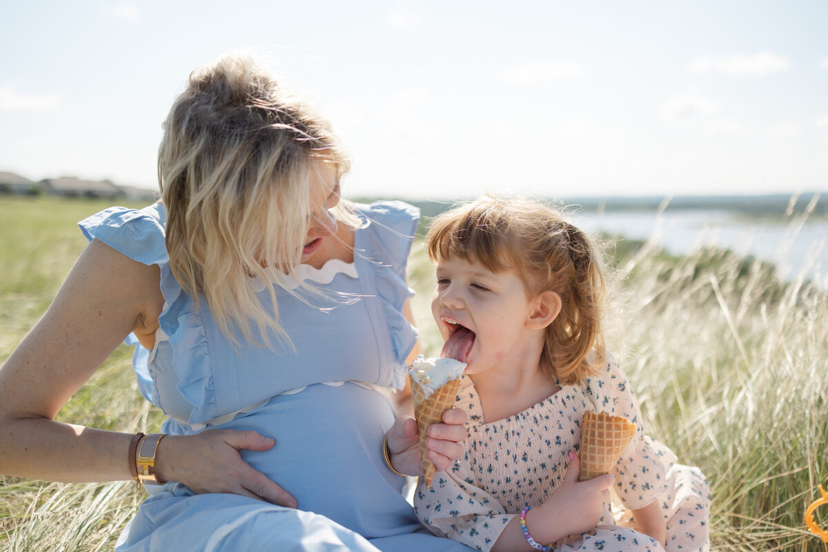 a pregnant mother in a blue dress holds out and ice cream cone for her daugter to lick as they sit in a soft grassy field with sunlight