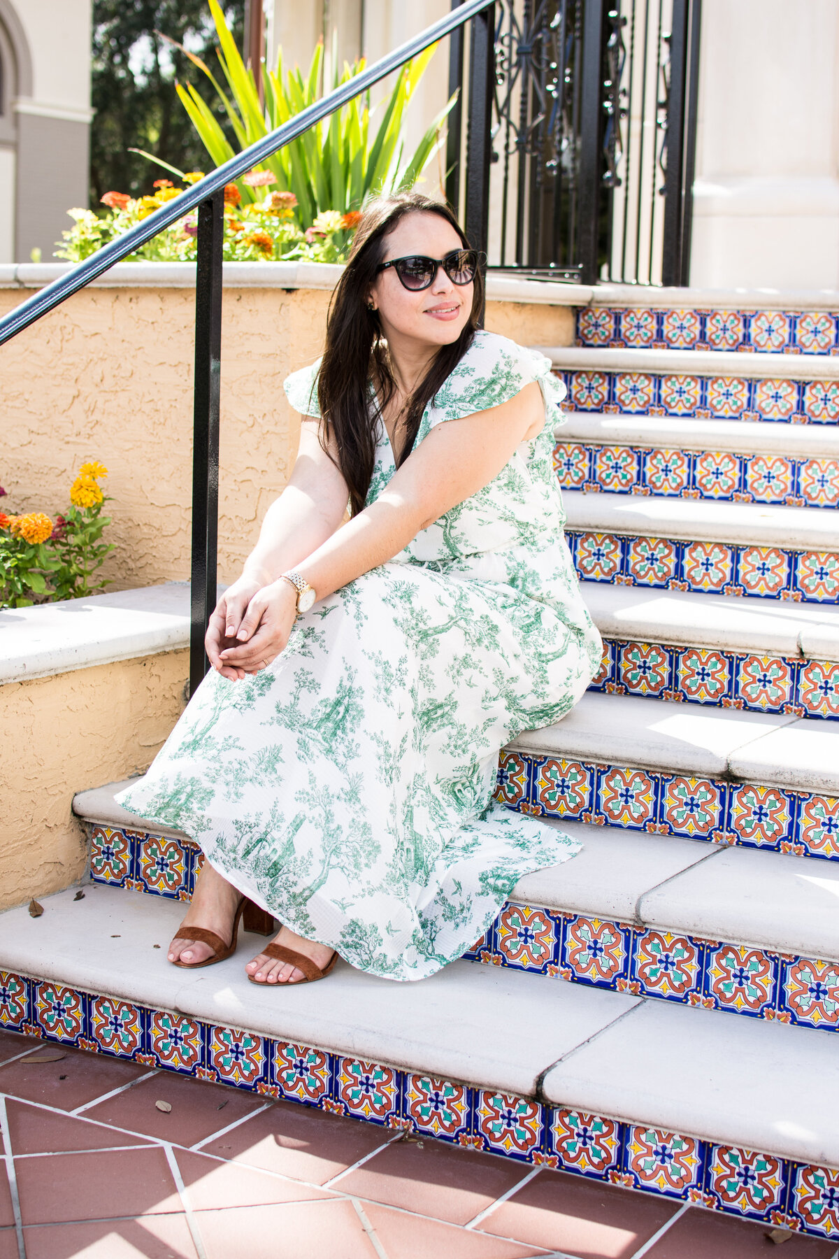 Woman with dark hair in white and green floral dress sits on stairs with decorative tiles