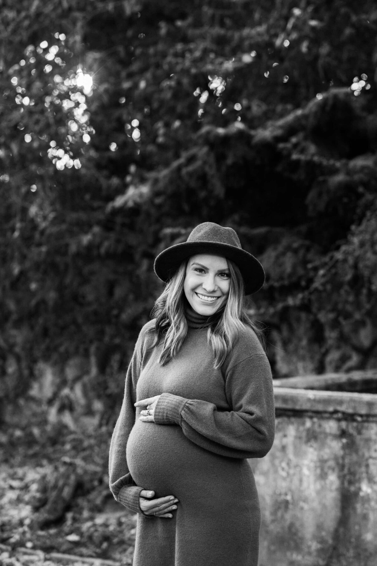 A smiling pregnant woman wearing a hat and holding her belly outdoors during her maternity photography session.