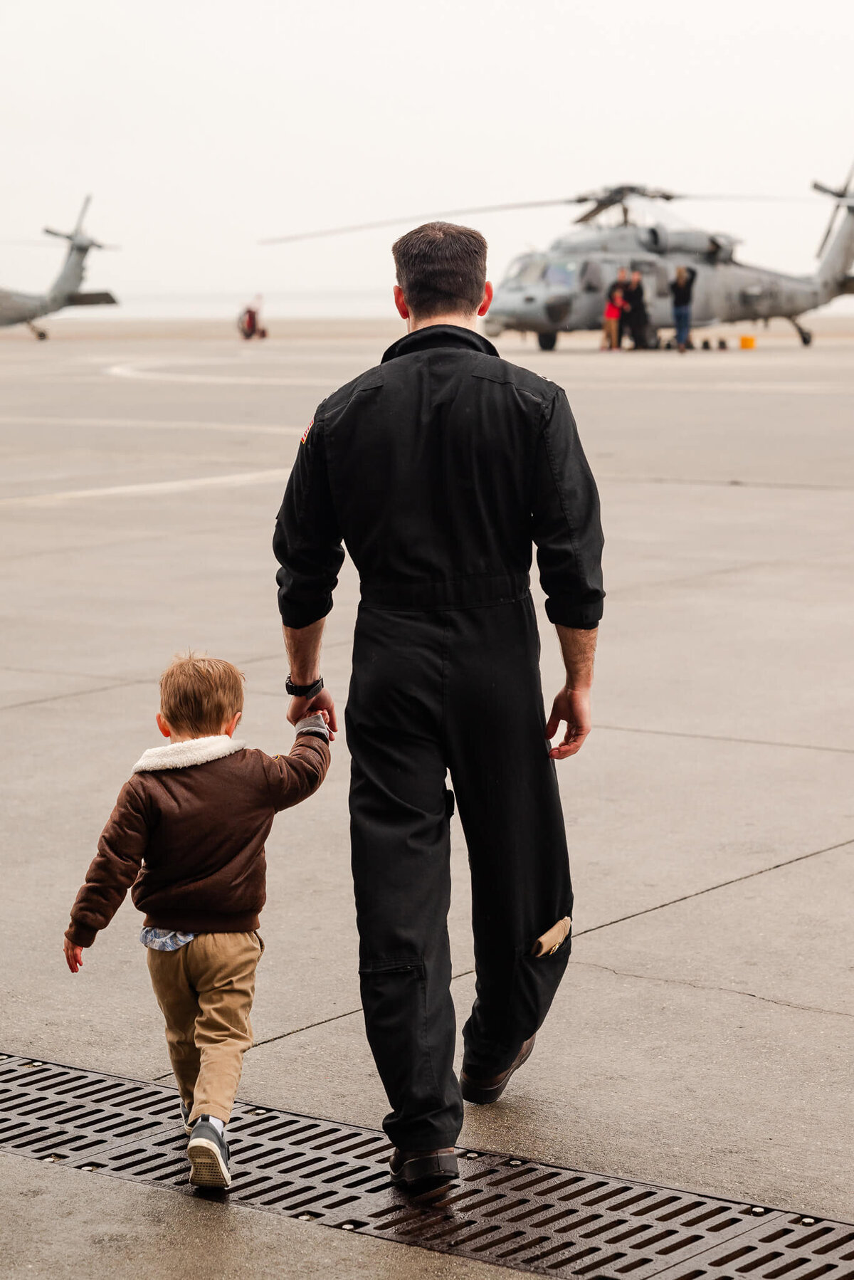 A young boy, wearing a flight jacket, holds his pilot dads hand as they walk to the helicopters during a military homecoming.