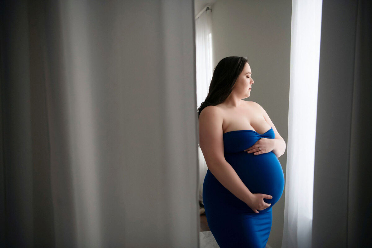 Pregnant woman in a blue strapless dress stands by a window, gently holding her belly. Soft light filters through white curtains, creating a serene, reflective mood.