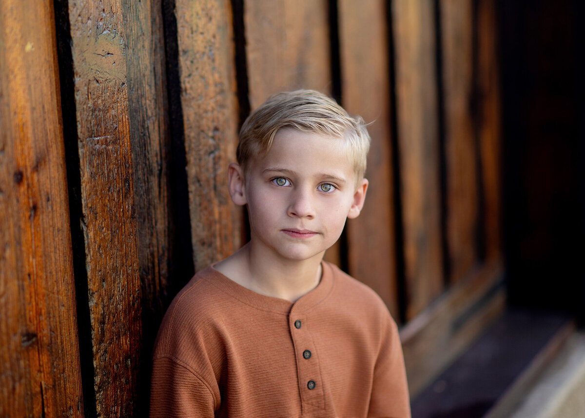 A child stands alone against a rustic wooden wall, their face serious