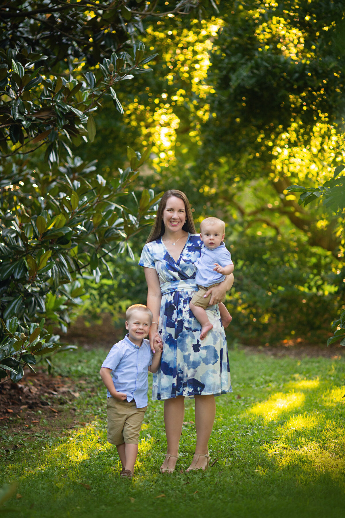A happy mother in a blue shirt walks through a park holding hands with her toddler son and carrying another for a New Jersey Family Photographer