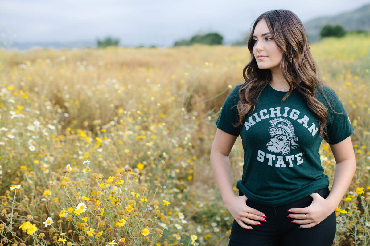 A woman stands in a field of yellow flowers, wearing a green Michigan State T-shirt. She has long brown hair and is looking to the right with her hands on her hips. The background is a blurred landscape.