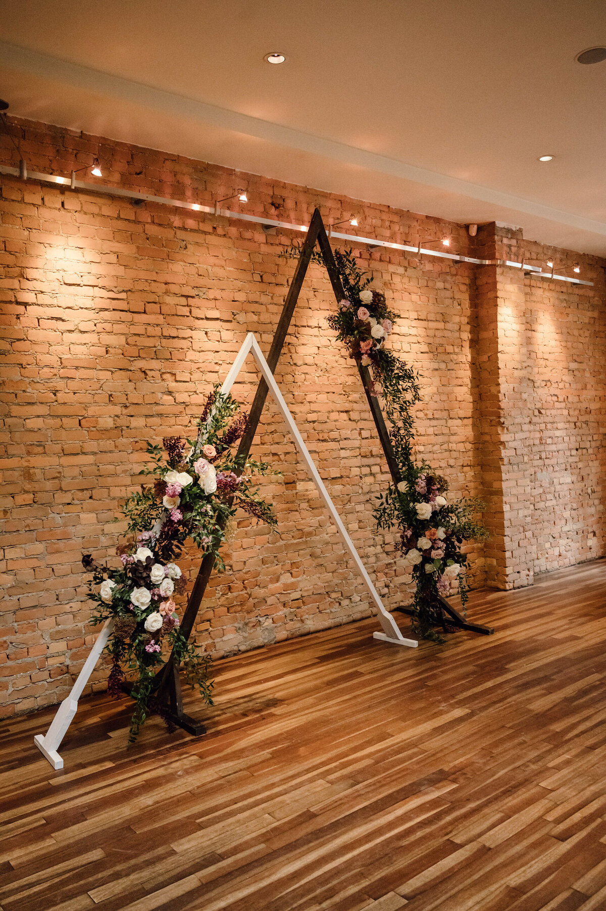 Indoor wedding ceremony with triangle arches and stunning white and burgundy florals at The Garret, historical and sophisticated, Calgary, Alberta wedding venue, featured on the Brontë Bride Vendor Guide.