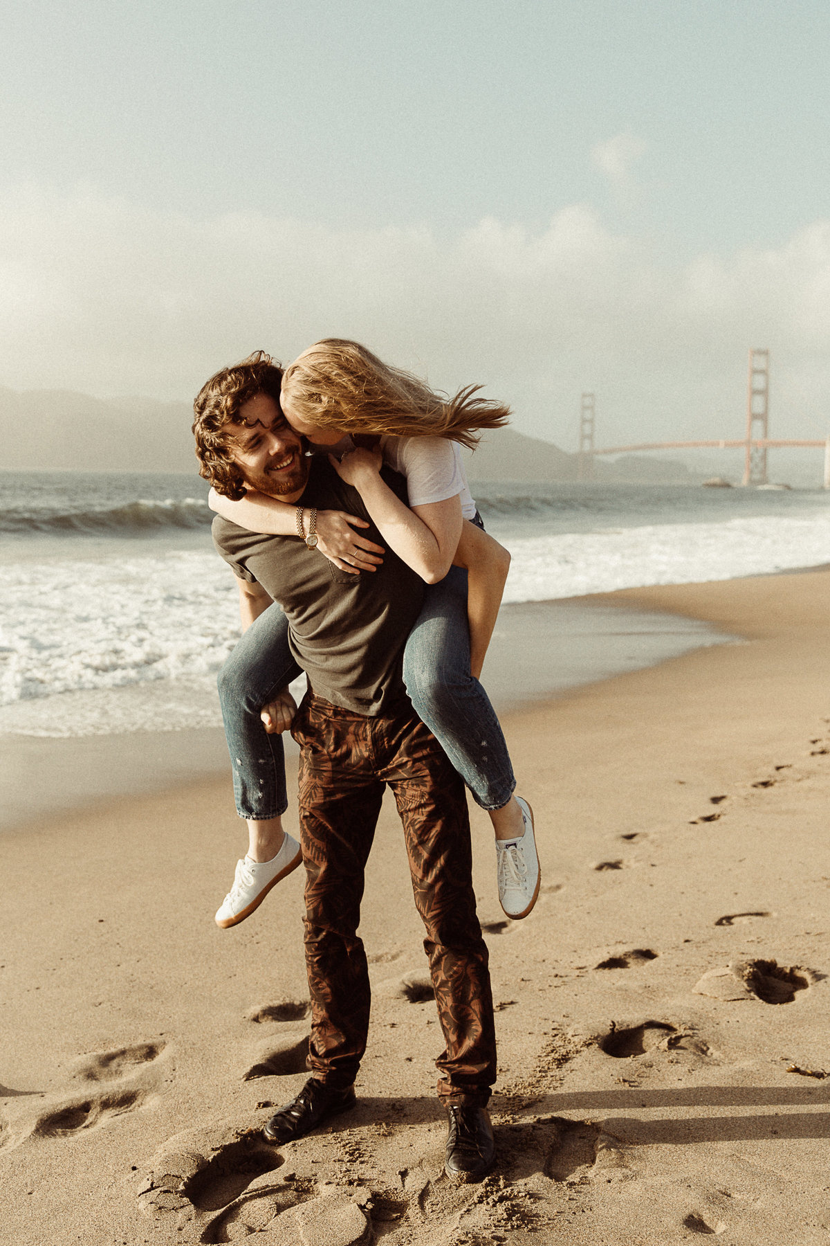 man gives woman a piggy back ride at Baker Beach with the Golden Gate bridge behind them