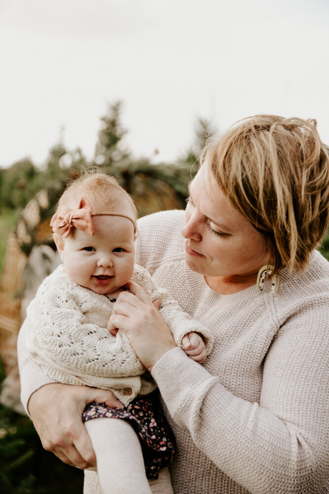 Mom is holding her infant daughter at Tree Lane Farms in London, Ontario for a top family Christmas photoshoot. The baby is smiling at the camera and the mom is smiling at the baby, touching her cheek.