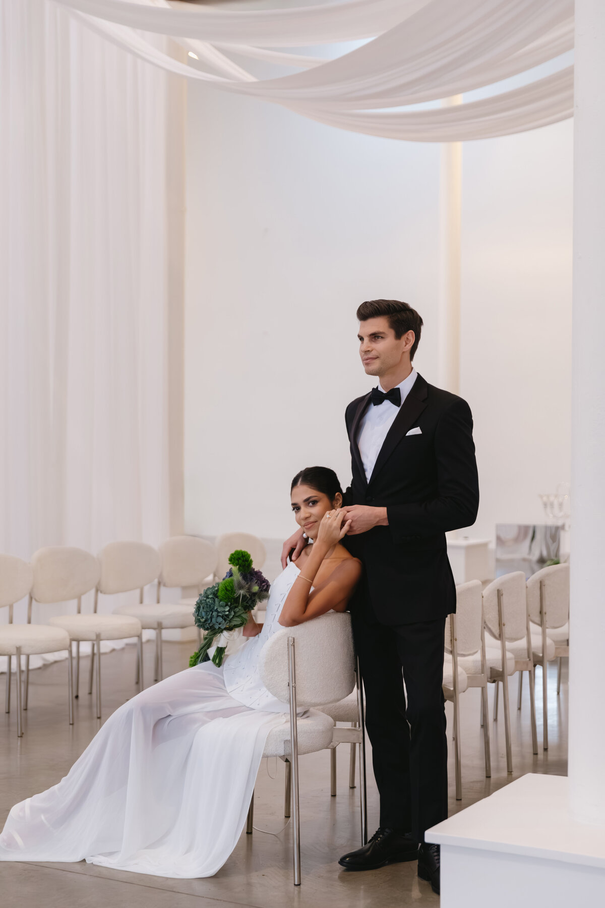Bride seated with groom standing protectively behind her in a modern, minimalist wedding editorial space.