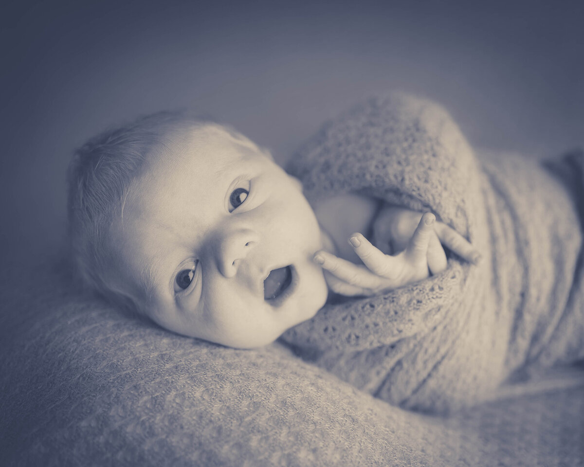 A newborn baby lays on a bed with mouth and eyes open in black and white