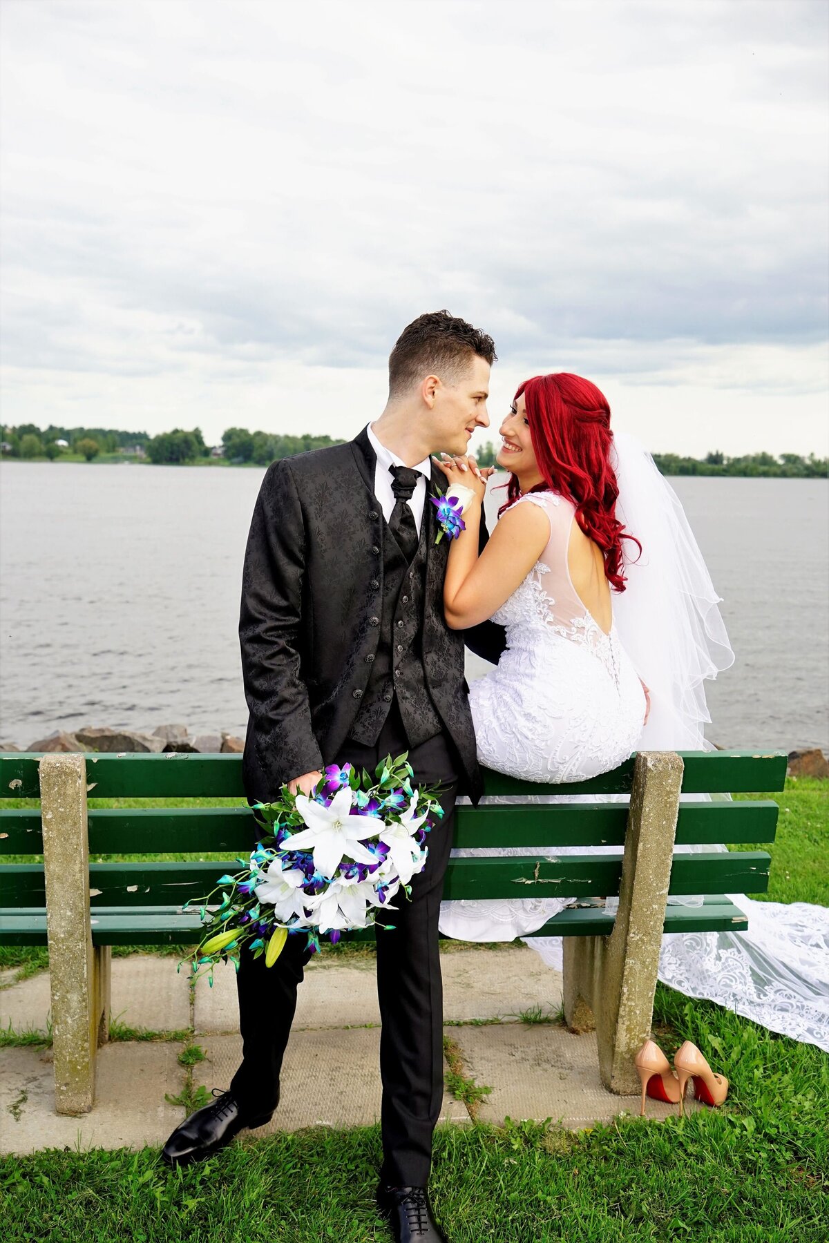 The bride with fiery red hair sits gracefully on a bench, gazing up at the groom who stands beside her. This intimate moment highlights the couple’s connection and the bride's striking hair color, set against a serene backdrop.