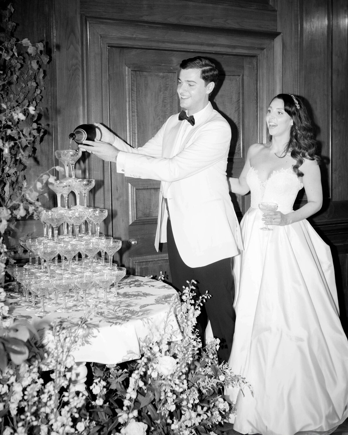 groom pours champagne into a champagne tower surrounded by meadows of flowers at luxury wedding at the ned london as his bride stands smiling next to him